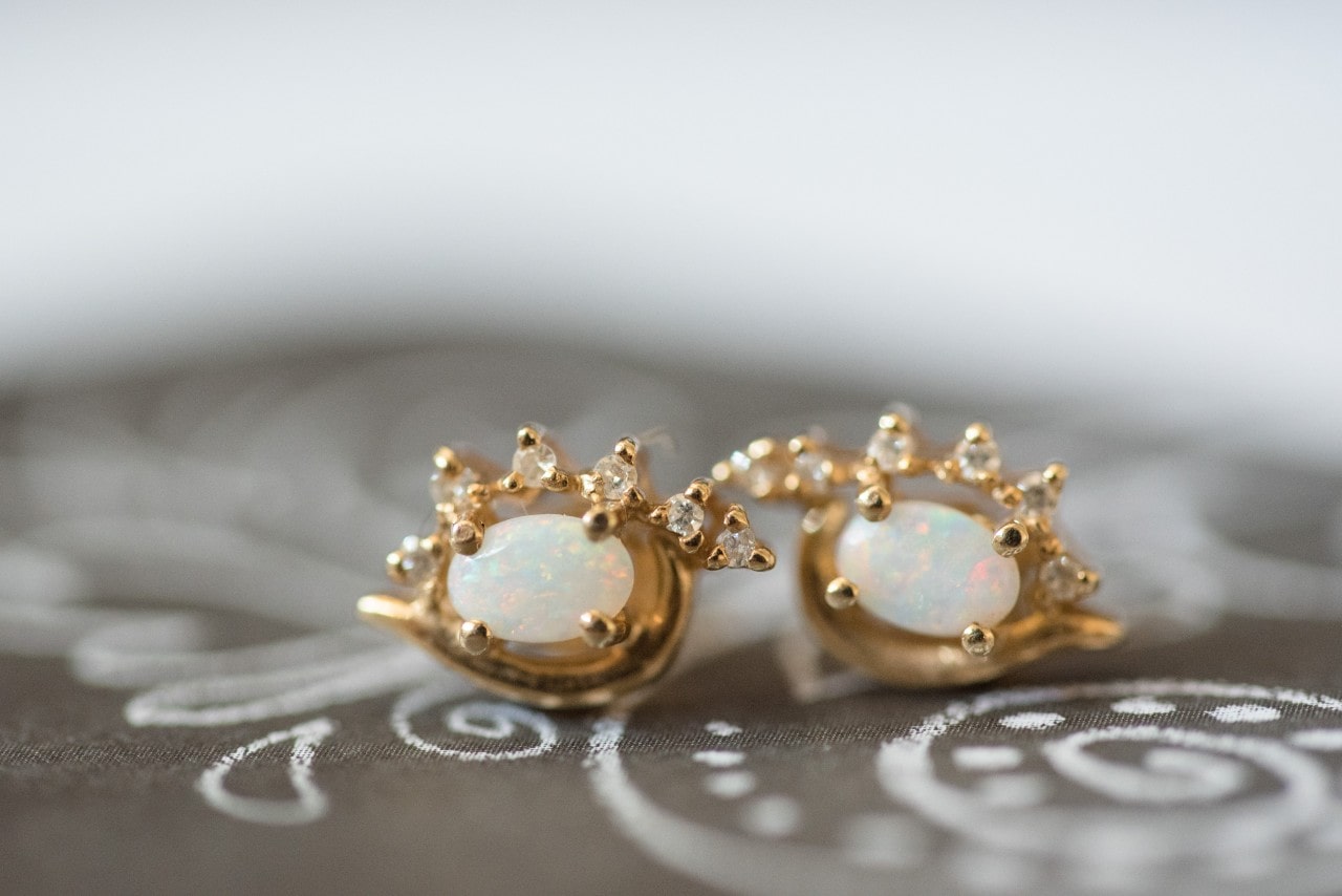 A closeup of a pair of yellow gold studs with prong-set opals and accenting diamonds sitting on a gray patterned fabric