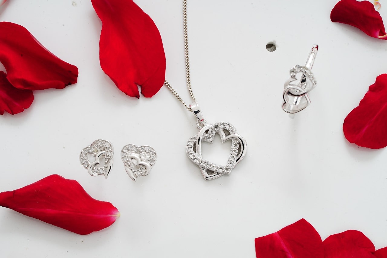 A closeup of a white gold overlapping heart accessory set, including stud earrings, a pendant, and a broach, on a white background with red rose petals