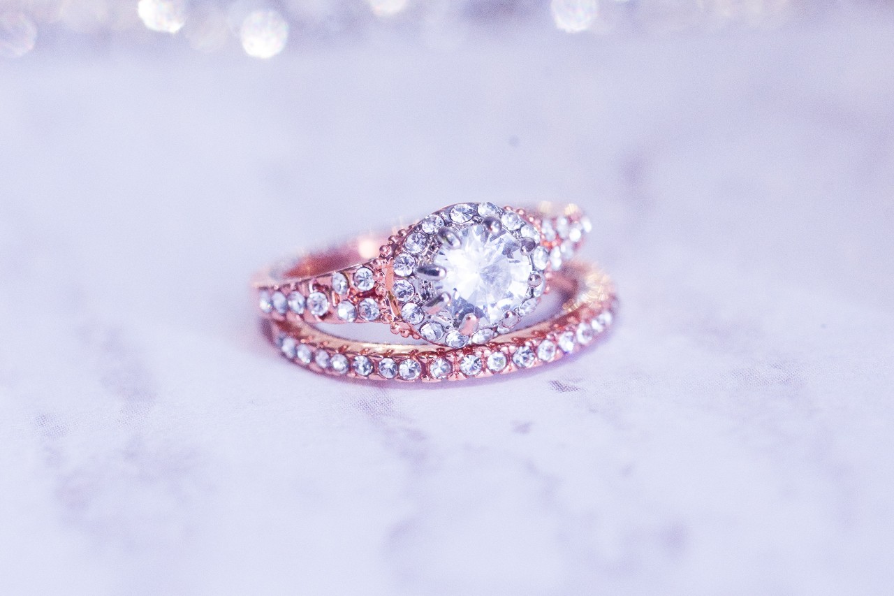 A vintage-inspired rose gold bridal set, including an eternity band and a round-cut halo engagement ring, sits on a marble countertop