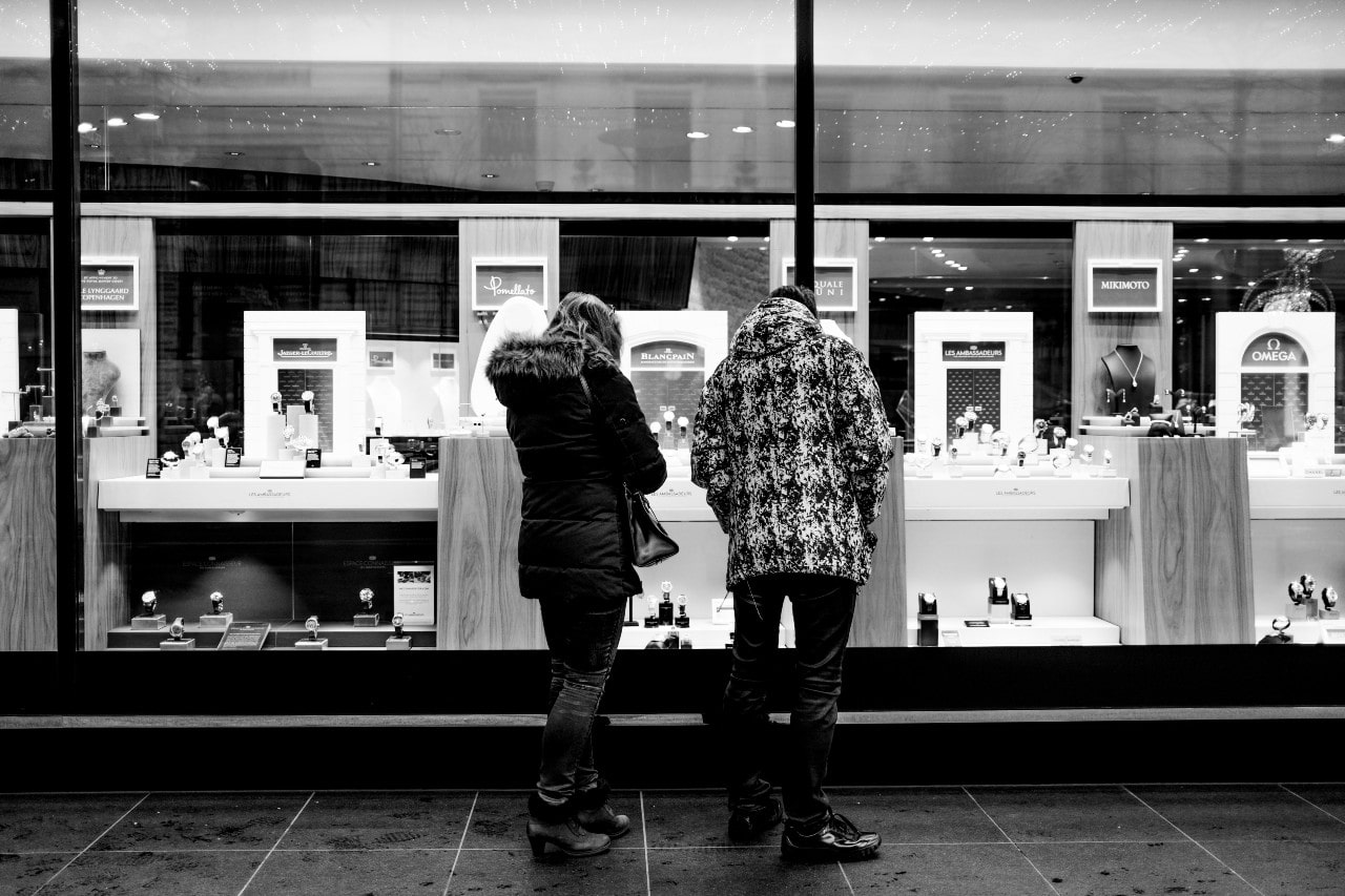 A couple browses the inventory of a closed jewelry shop through the window while on an evening stroll in the city