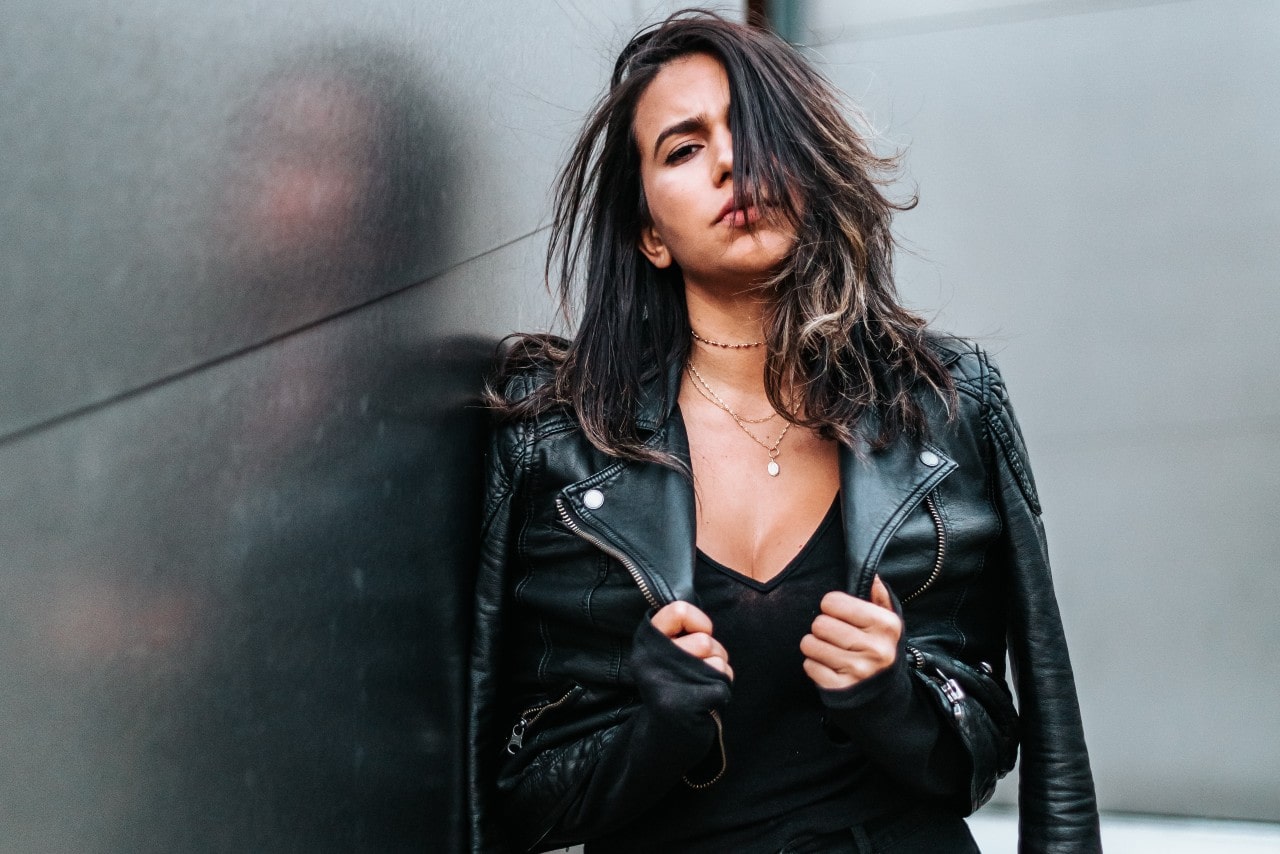 A woman next to a building wearing a black leather jacket, black v-neck, and multiple chain necklaces with one pendant layered together