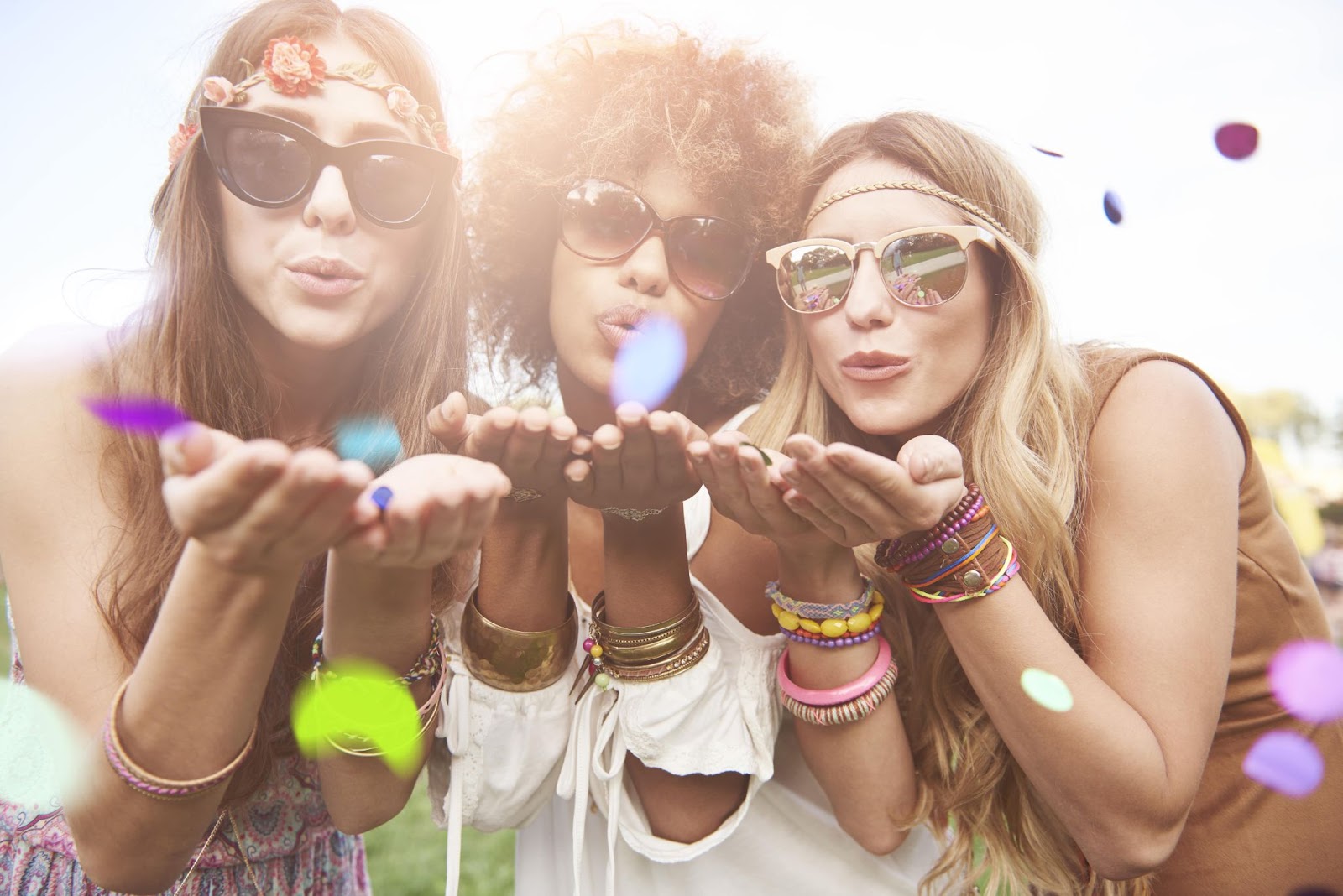 Three friends blowing confetti into the camera, smiles on their faces and many bracelets on wrists along with sunglasses on each of them