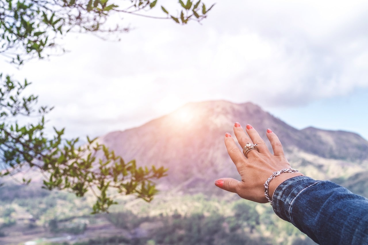 A woman on a morning hike reaches out toward the rising sun as she wears her bridal jewelry and bracelet